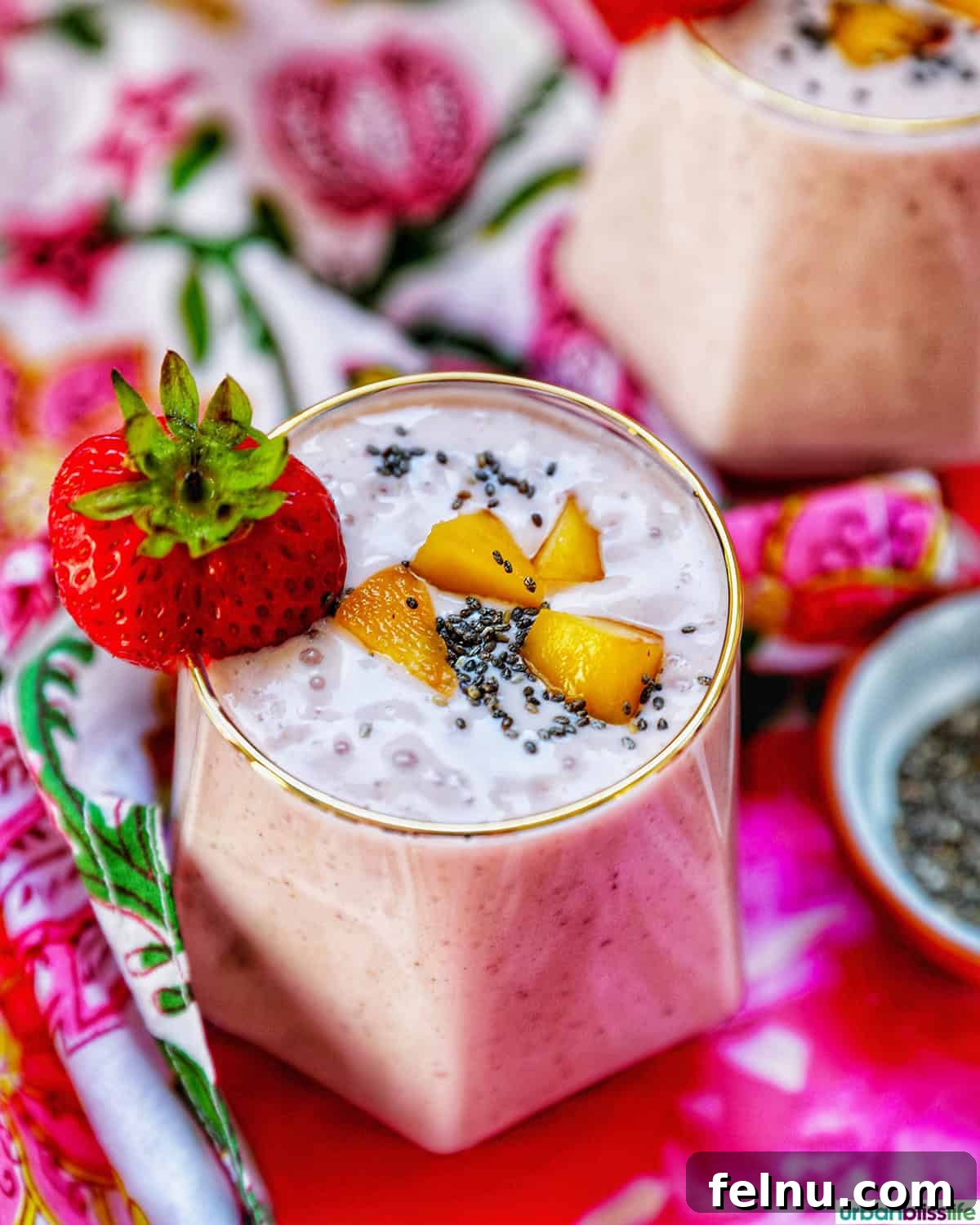 Peachy Berry Bliss 11 A glass of strawberry peach smoothie, beautifully garnished with a fresh strawberry, set against a backdrop of colorful napkins and a second smoothie.