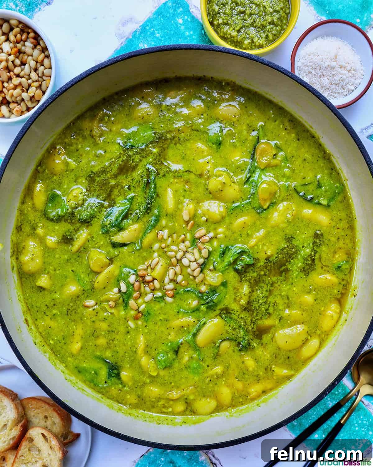 large pan of pesto beans with side bowls of pine nuts, parmesan cheese pesto sauce, and crostini slices.