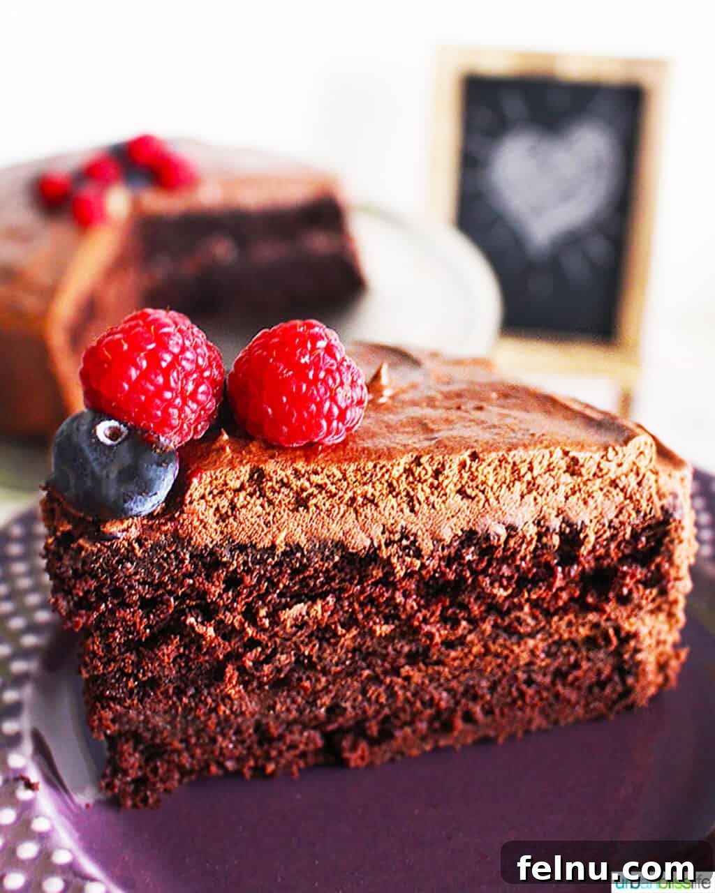 Slice of vegan chocolate cake with chocolate buttercream frosting and fresh fruit on a purple plate.