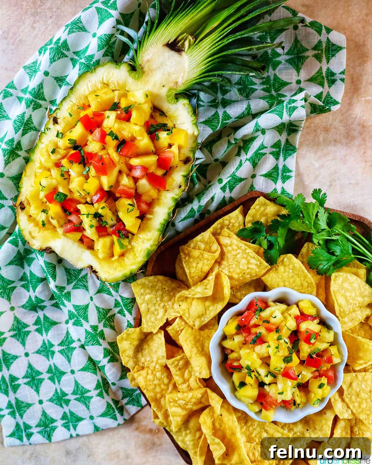 Sun-Kissed Fruit Salsa 7 A decorative display of pineapple mango salsa, with a small bowl of salsa and tortilla chips beside it, all nestled within a hollowed-out pineapple shell, resting on a green and white patterned napkin.