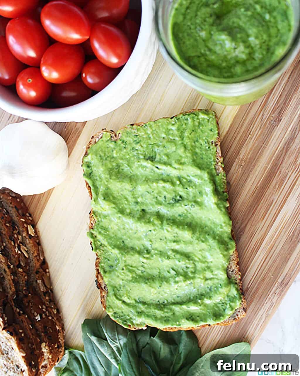Creamy Avocado Pesto 3 avocado pesto spread on a piece of bread, with jars of tomatoes, pesto, cheese, and spinach leaves places around the bread on a cutting board.