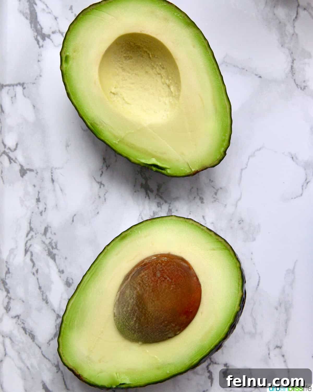 Creamy Avocado Pesto 4 Avocado split in half with flesh and pit facing up, on a marble countertop.
