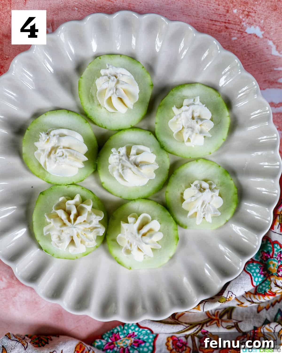 Crisp Cucumber Shrimp Delights 9 Multiple cucumber slices, each topped with a delicate swirl of cream cheese, arranged on a scalloped plate against a soft pink background.