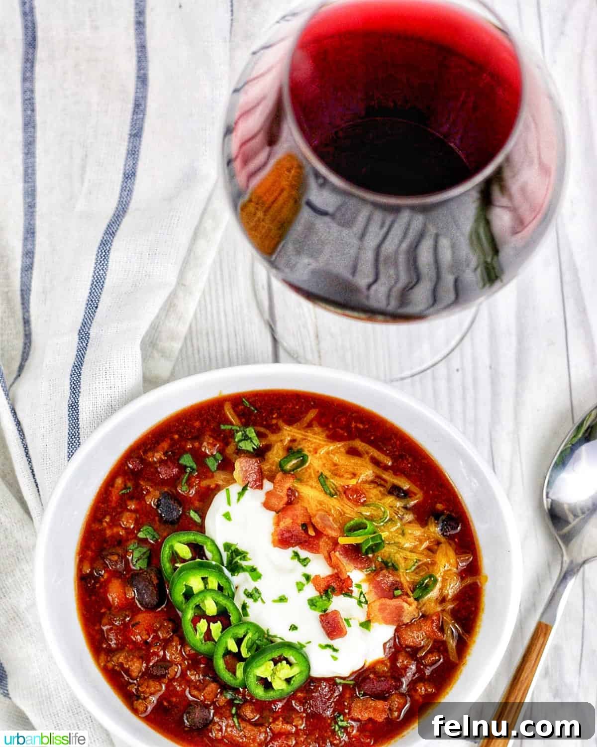 Hearty & Healthy Turkey Chili (Instant Pot Friendly!) 5 A beautifully styled bowl of chili, topped with jalapenos, sour cream, crispy bacon, and shredded cheese, accompanied by a glass of red wine on a white table with a spoon.