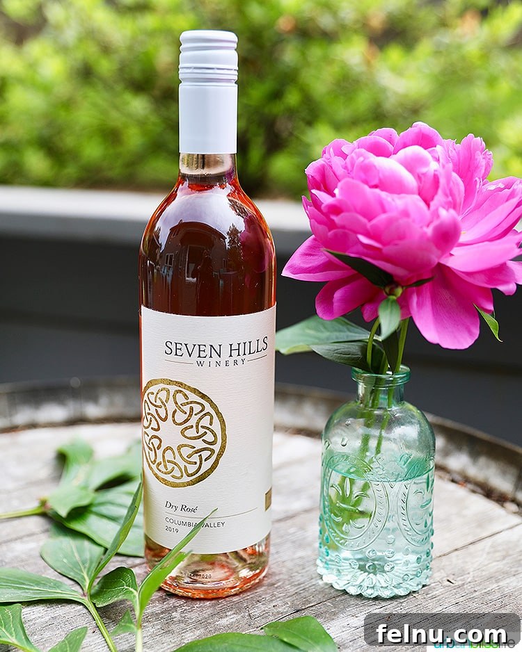 Toast to Summer: 12 Perfect Rosés for National Rosé Day 10 A bottle of Seven Hills Winery 2019 Rosé, with a delicate pink flower in a vase beside it, suggesting elegance and freshness.