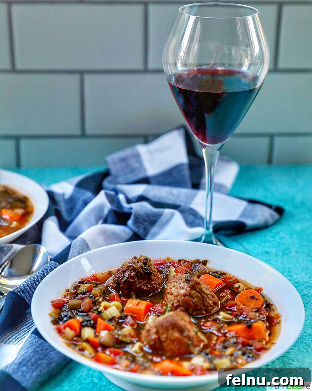 Hearty Slow Cooker Meatball Soup 5 A glass of full-bodied red wine next to a steaming bowl of meatball soup on a blue checkered tablecloth.