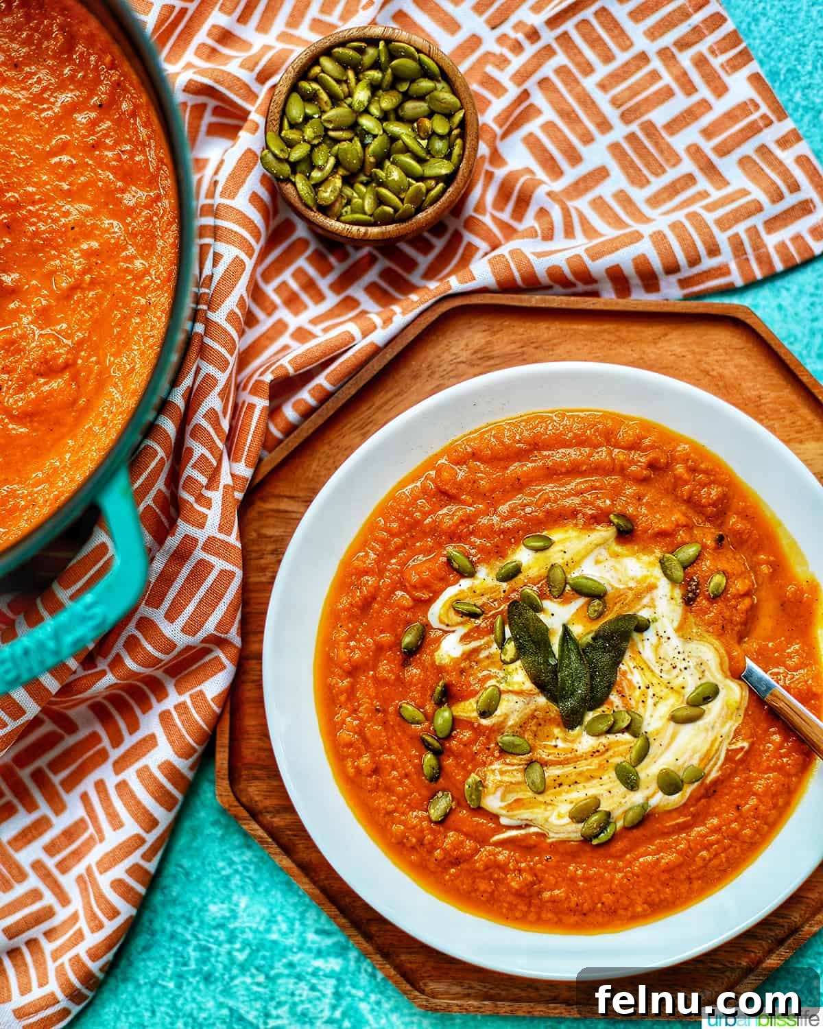 A pristine white bowl of vegan pumpkin soup sits on a rustic wooden tray, next to a Dutch oven filled with more soup, set against a vibrant blue background with an orange and white napkin.