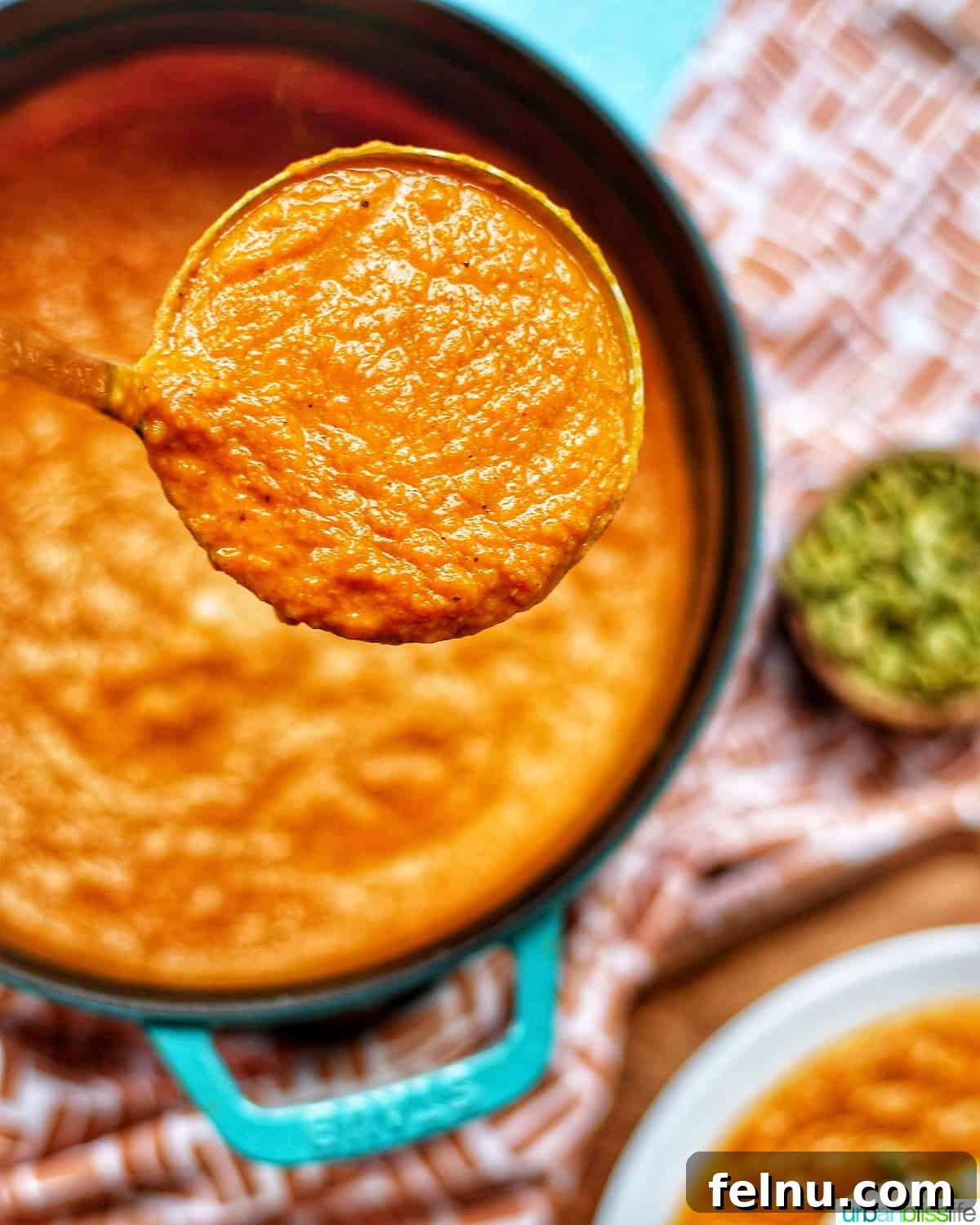 A ladle full of vibrant vegan pumpkin soup without cream, lifted from a Dutch oven, with additional soup bowls and pumpkin seeds in the soft-focus background.
