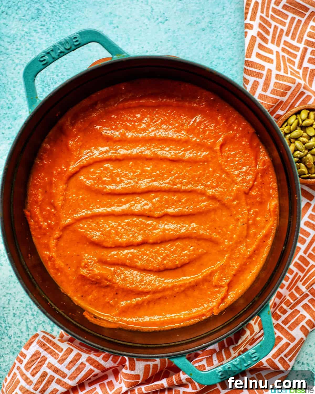 A bright orange vegan pumpkin soup simmering in a vibrant red Staub Dutch oven, with a small bowl of pumpkin seeds on the side, against a cheerful blue background.