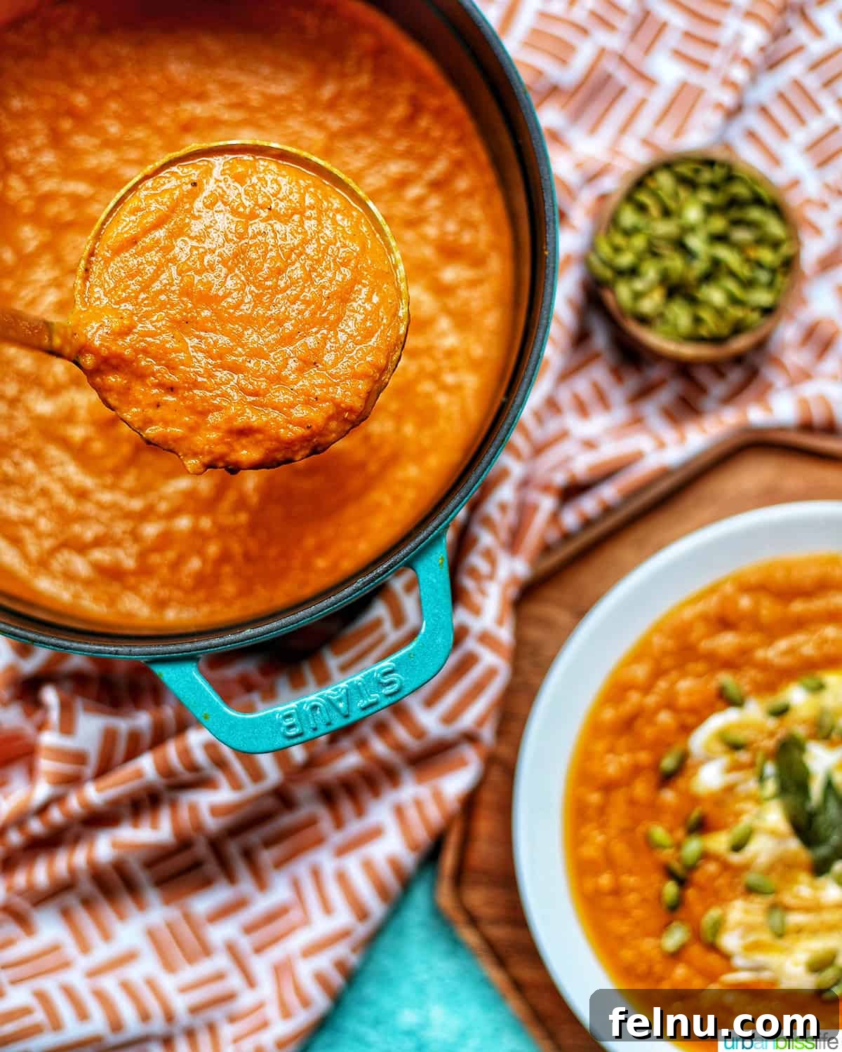 A ladle of rich, bright orange vegan pumpkin soup held above a Dutch oven brimming with the creamy soup, with bowls and pumpkin seeds in the background.