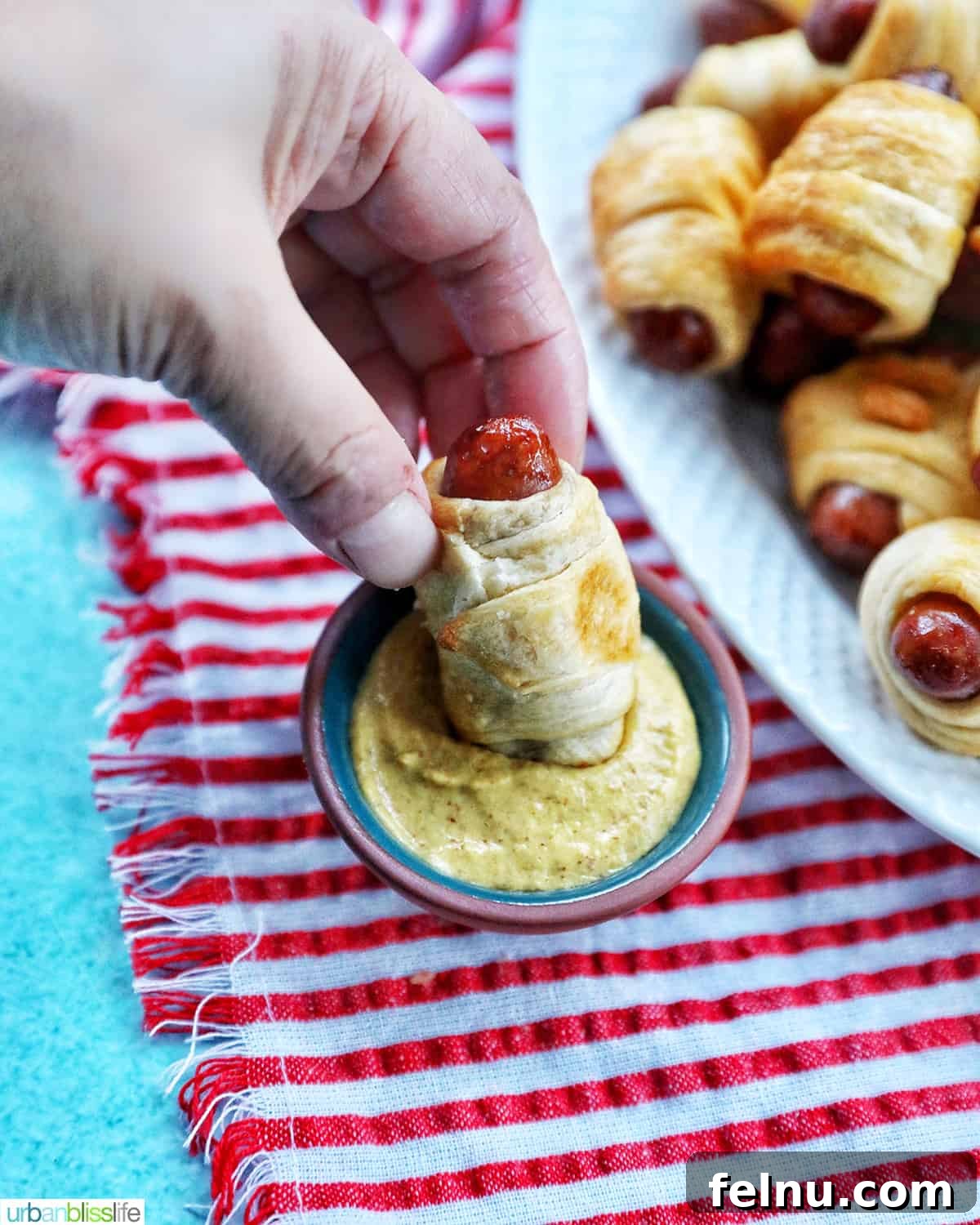 Crispy Air-Fried Pigs in a Blanket 14 a hand dipping Air Fryer Pigs in a Blanket into mustard on a red and white napkin with several pigs-in-a-blanket on a plate.