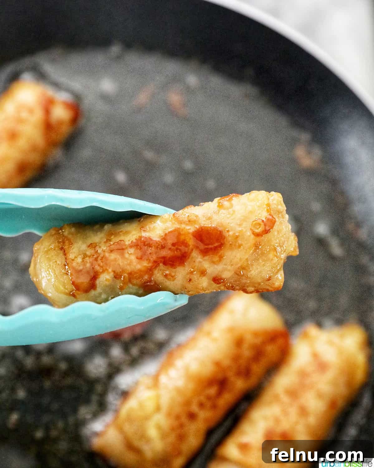 Effortless Filipino Turon: Crispy Banana Lumpia Perfection 24 Tongs holding up a fried banana turon lumpia with more Filipino turon frying in the background.