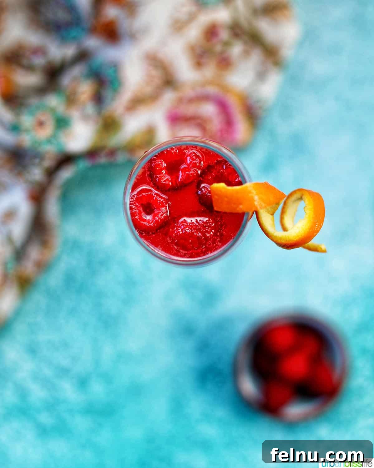 Raspberry Sparklers 3 Overhead view of a Raspberry Mimosa in a champagne flute, garnished with raspberries and a twist of orange rind, against a vibrant blue backdrop.