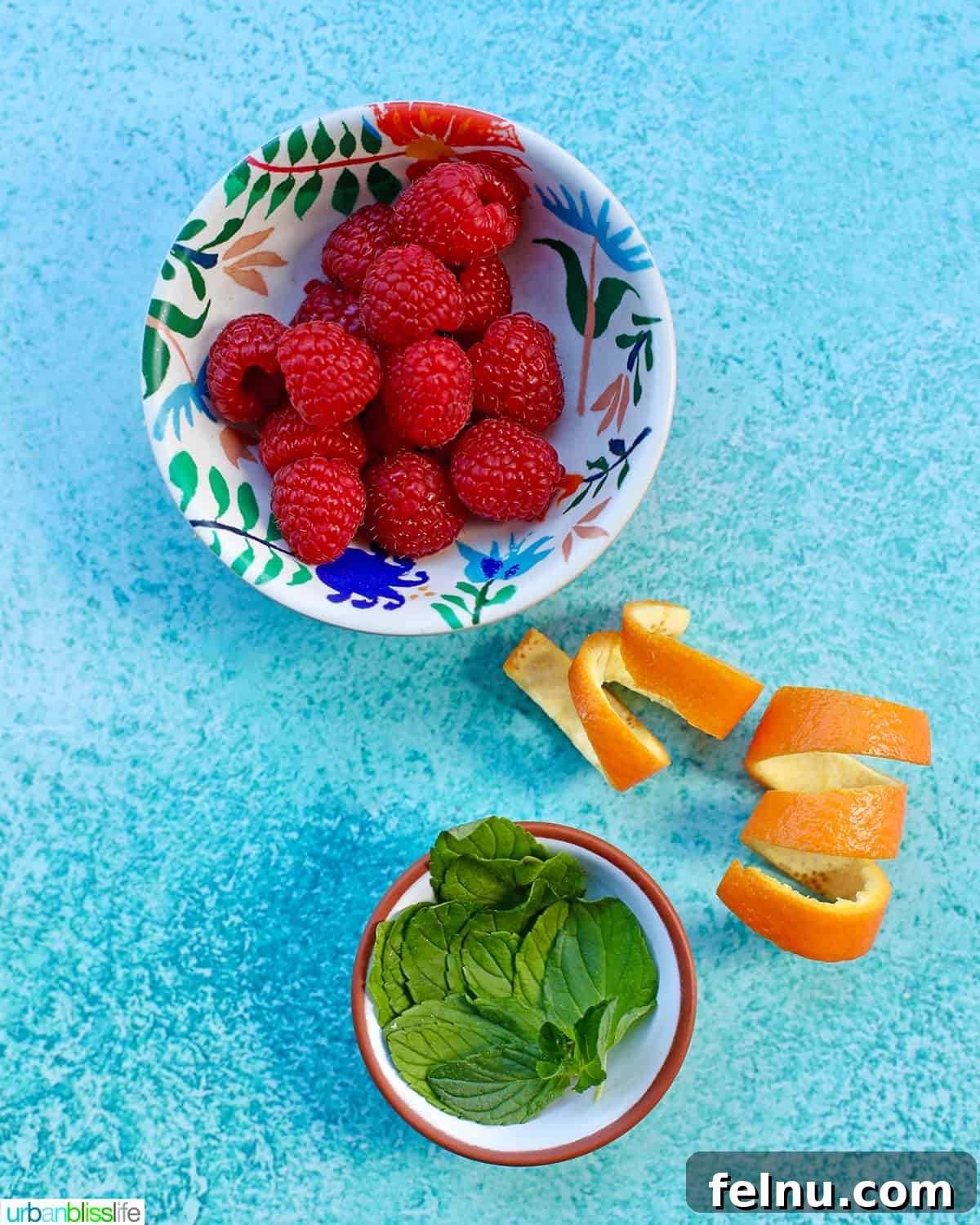 Raspberry Sparklers 5 Bowls containing raspberries, fresh mint leaves, and orange rind, prepared as garnishes for raspberry mimosas, all displayed on a blue surface.