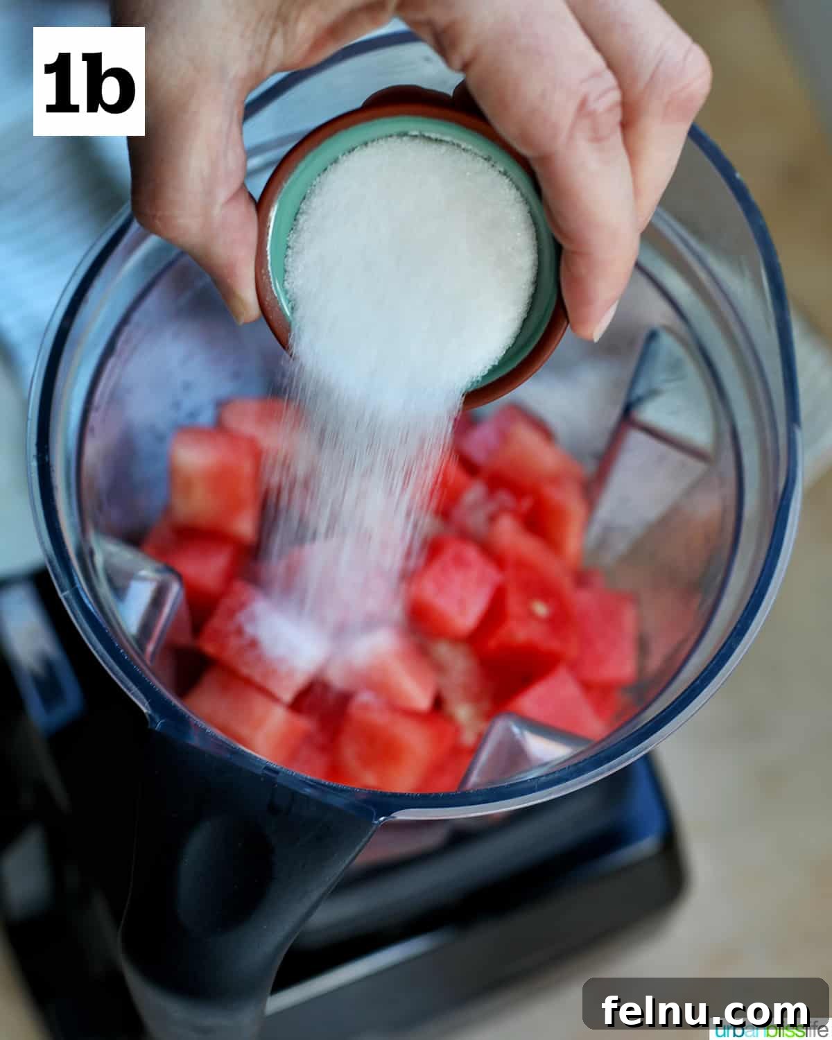 Watermelon Sunset Fizz 7 A hand adding granulated sugar from a small bowl into a blender containing cubed watermelons, ready to be blended into a sweet and smooth mimosa mixture.