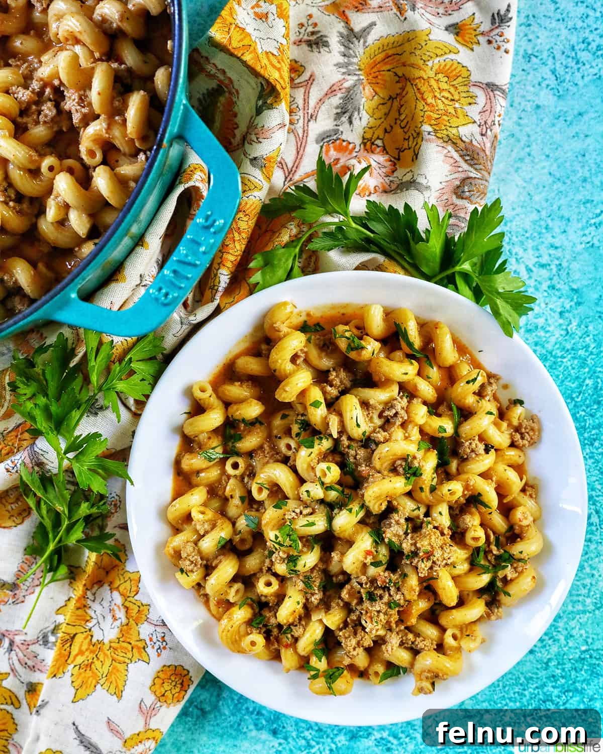 Creamy Dairy-Free Cheeseburger Mac Skillet 13 A white bowl filled with dairy-free cheeseburger pasta, with a fork, placed on a blue background, ready to be enjoyed.