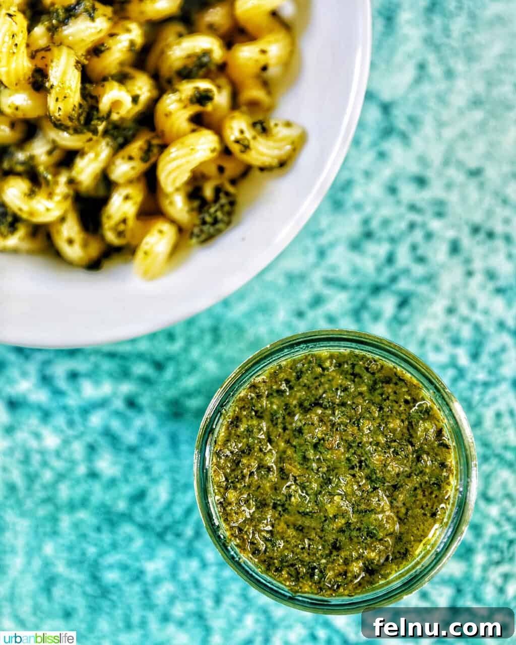 Zingy Mint Pesto 11 A glass jar of fresh mint pesto in the foreground, with a bowl of pesto pasta artfully arranged in the top left, all presented on a vibrant blue background.