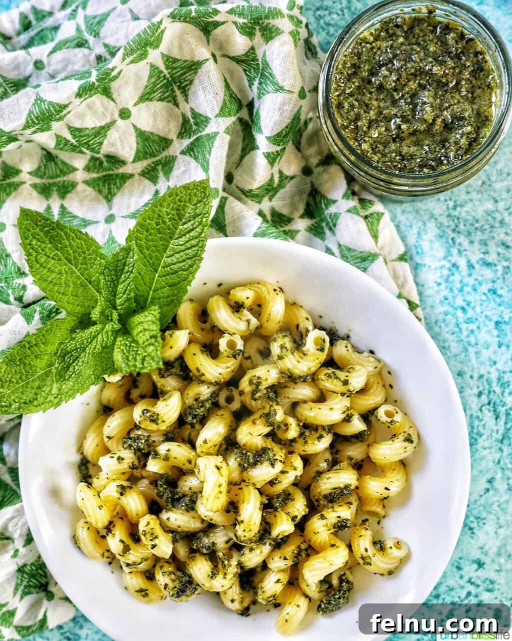 Zingy Mint Pesto 13 A pristine white bowl showcasing pasta coated in vibrant mint pesto, adorned with fresh mint leaves, positioned alongside a jar of mint pesto. The setting is a bright blue background with a green and white napkin to the left.