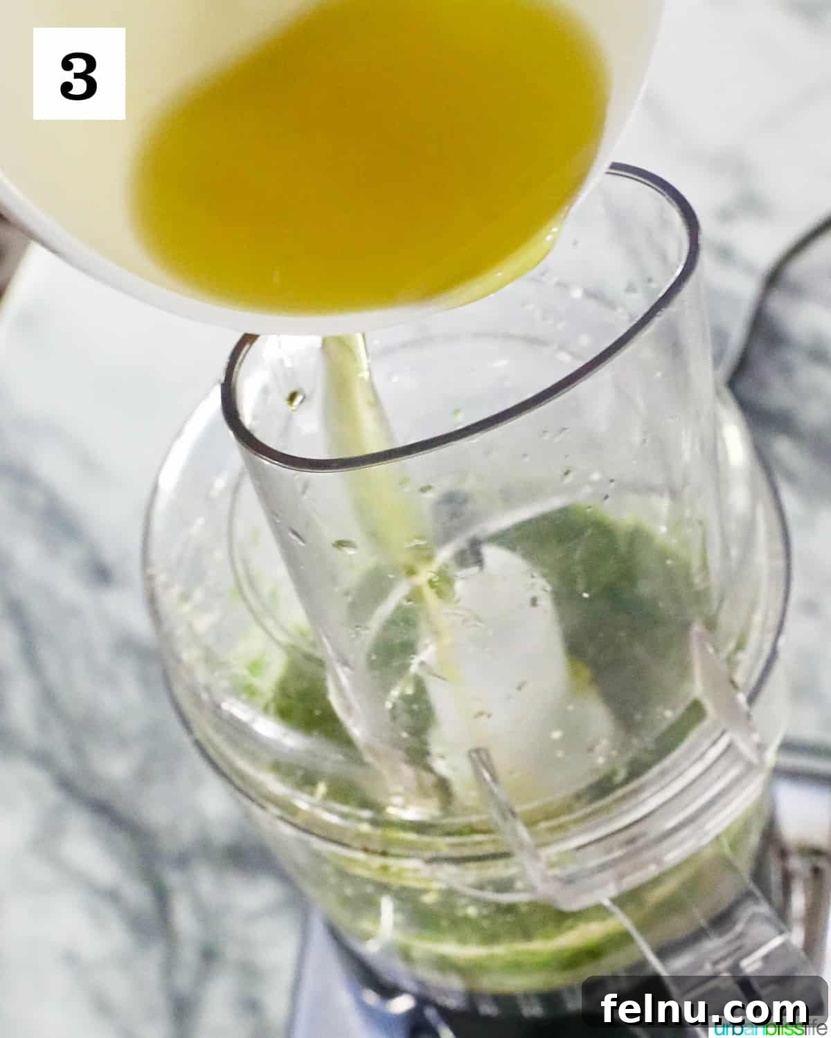Zingy Mint Pesto 9 A stream of golden olive oil being poured from a white bowl into the top opening of a food processor, where mint pesto ingredients are actively blending.