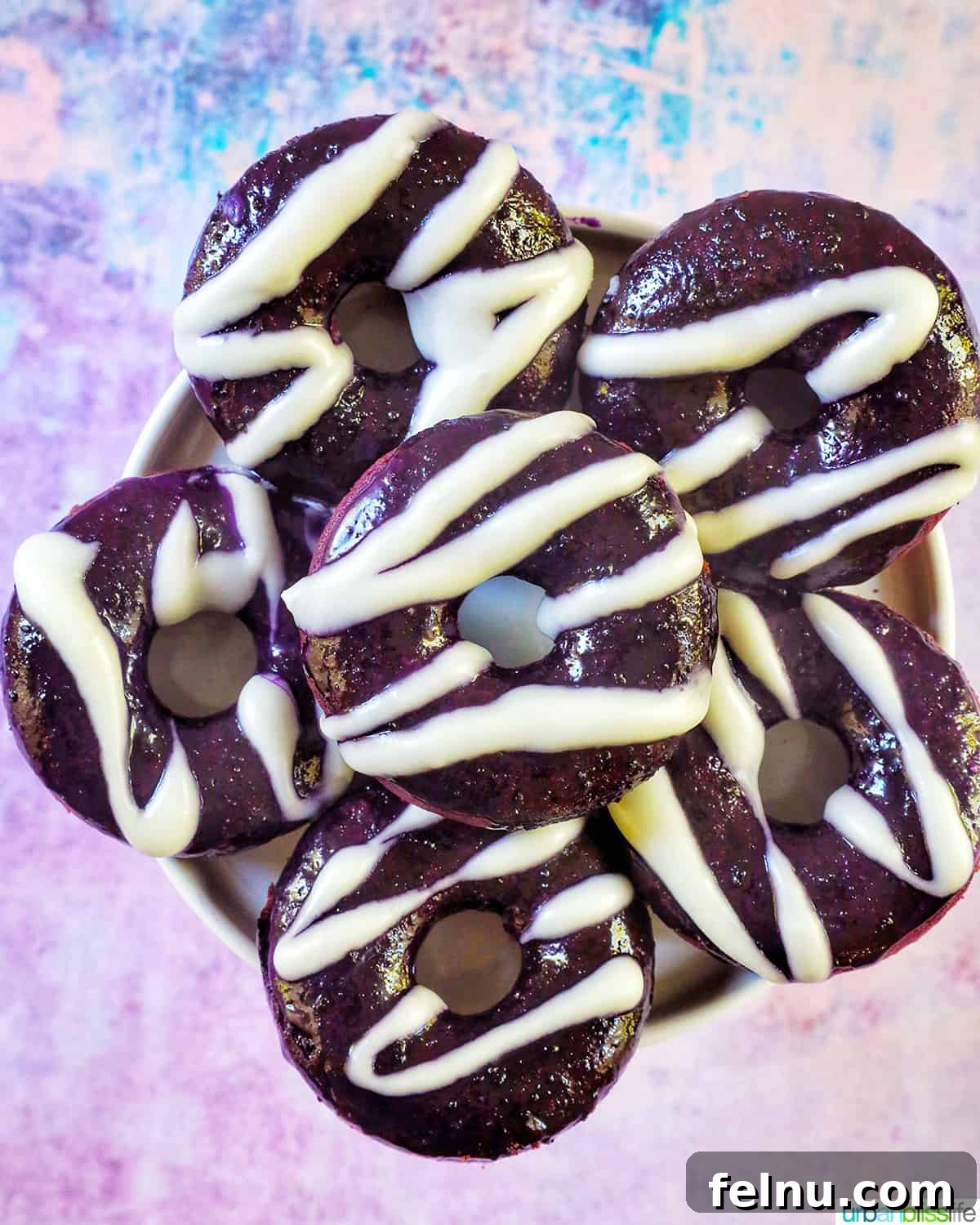 Ube Dream Donuts 2 Several ube donuts adorned with ube and white glaze, displayed against a vibrant purple and blue backdrop.