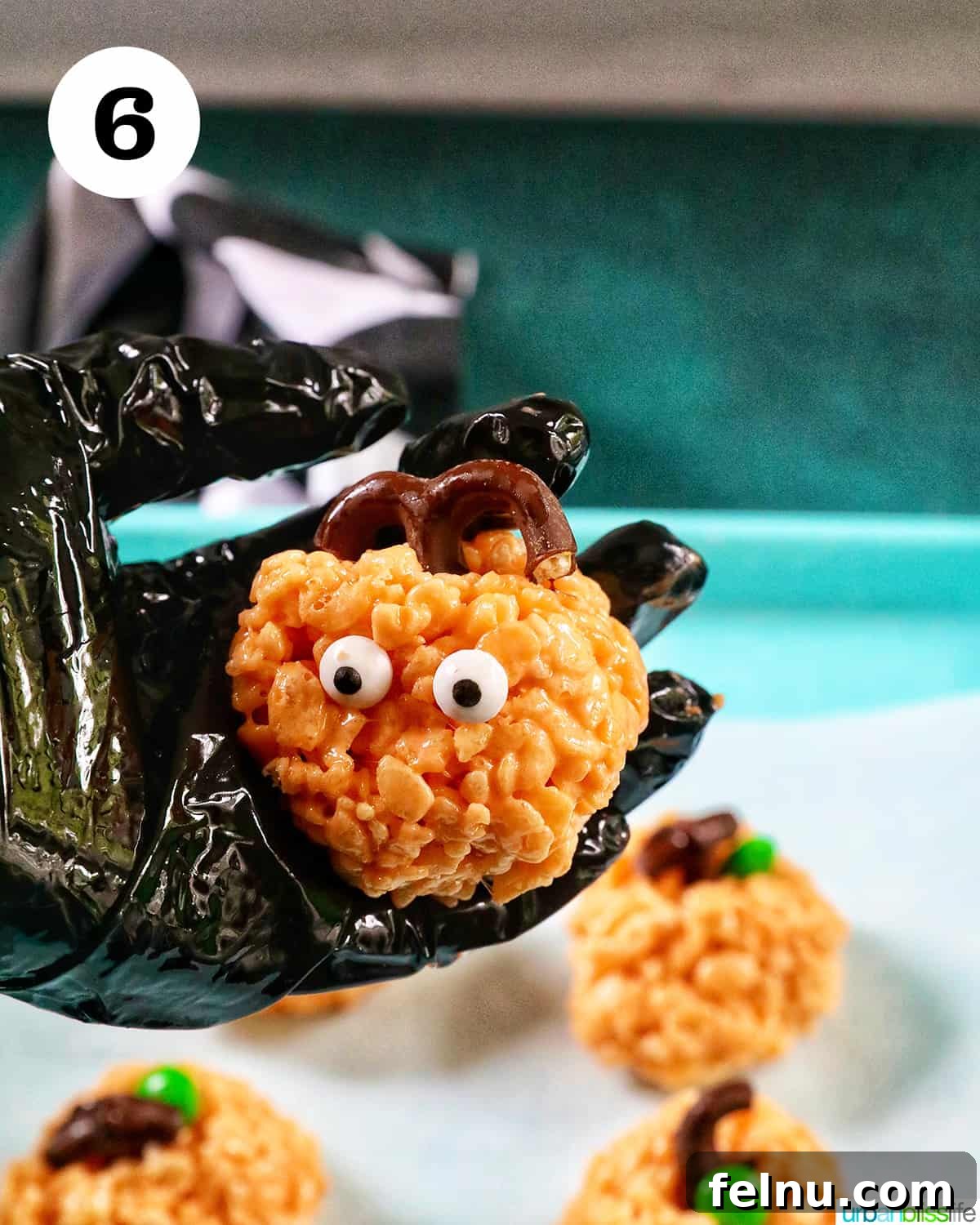 Seasonal Pumpkin Crunch Squares 13 A gloved hand holding a decorated pumpkin rice krispie treat, showing the addition of a pretzel stem and green candy leaf.