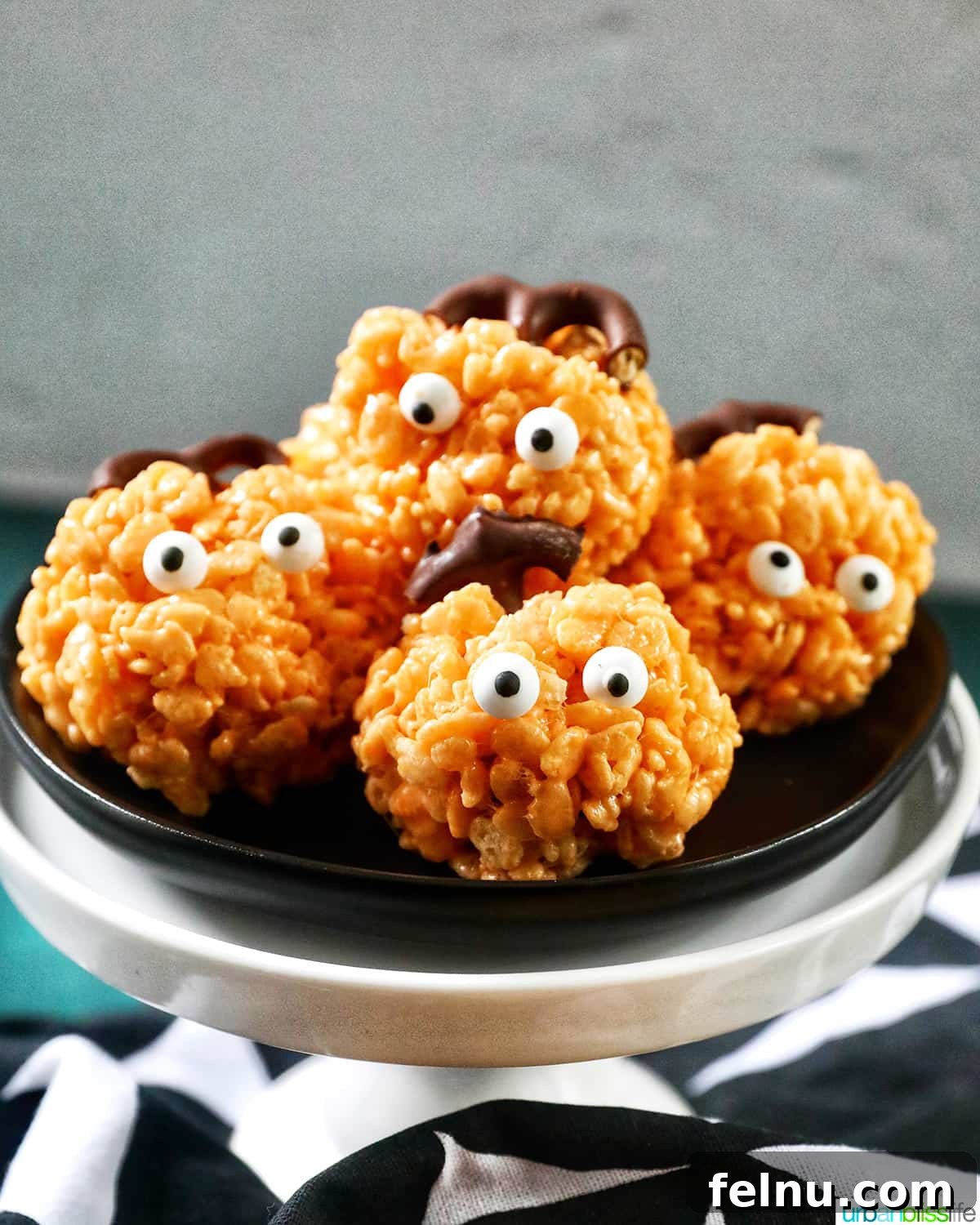 Seasonal Pumpkin Crunch Squares 16 Two orange pumpkin rice krispie treats with candy eyes, one with a pretzel stem and green leaf, on a black plate.