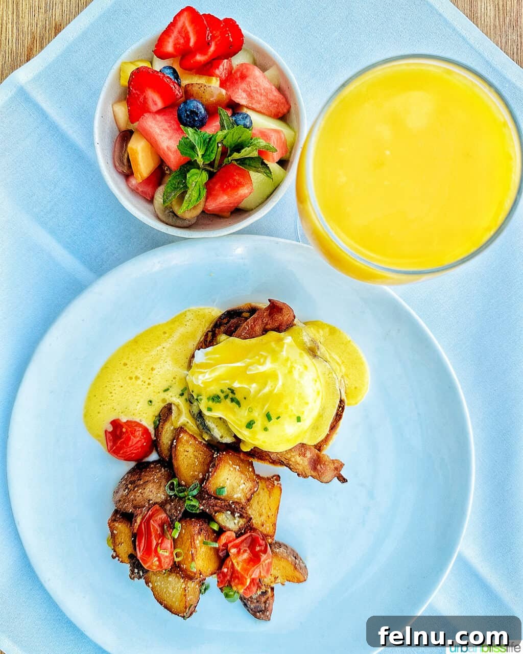 The Laurel Point Haven 8 eggs benedict with potatoes on a plate, with glass of orange juice and side of fruit.