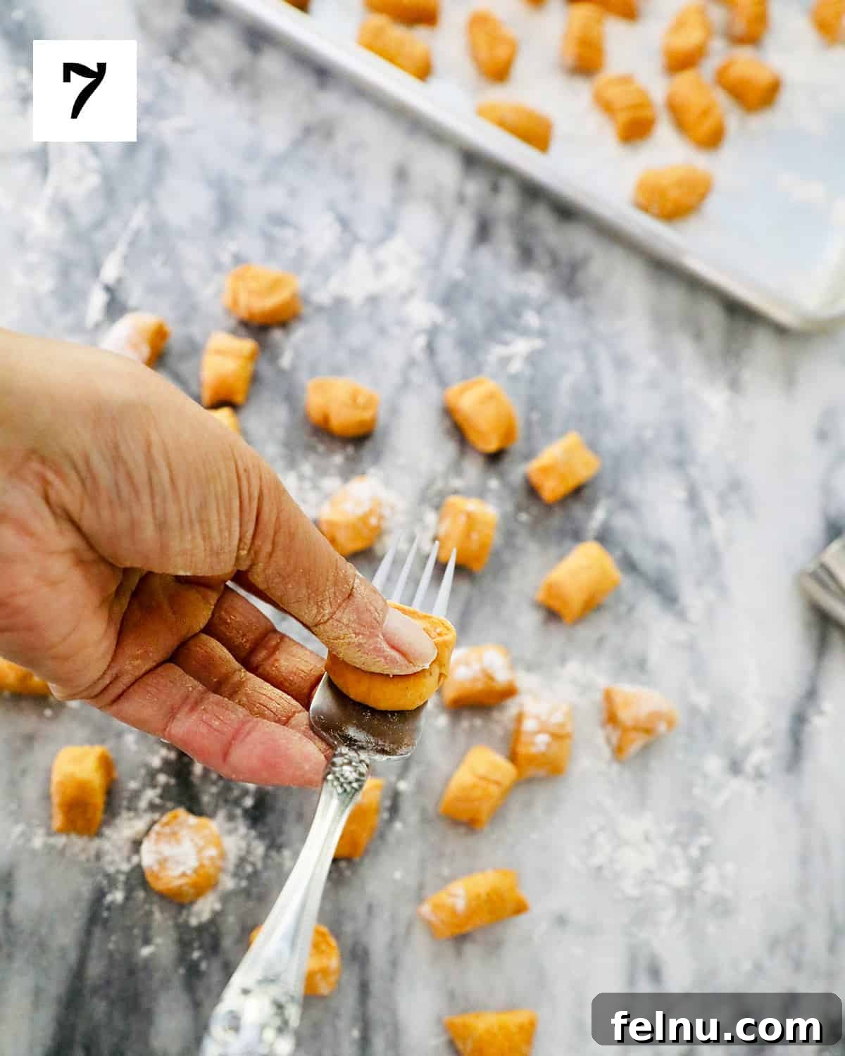 A hand expertly shapes a piece of pumpkin gnocchi by pressing and rolling it against the tines of a fork on a marble countertop, creating the characteristic ridges.