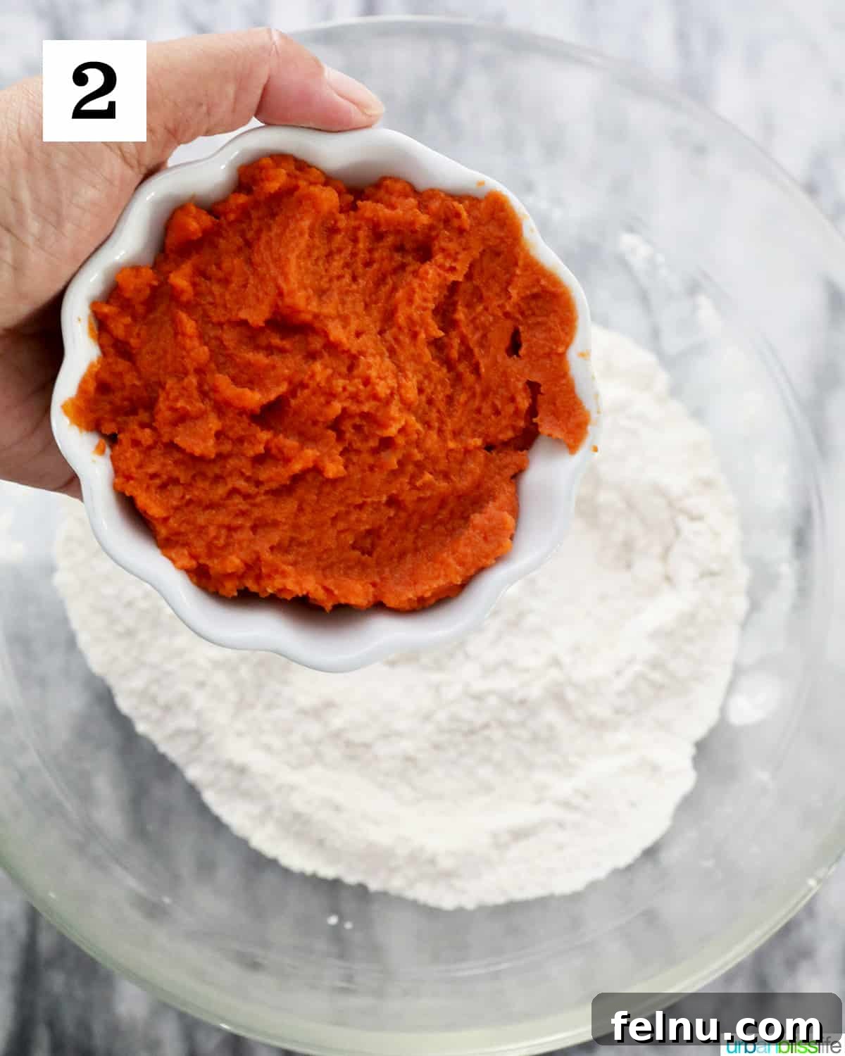 A bowl of vibrant pumpkin puree is carefully added to a mixture of dry ingredients in a larger bowl on a marble countertop, initiating the pumpkin gnocchi dough preparation.