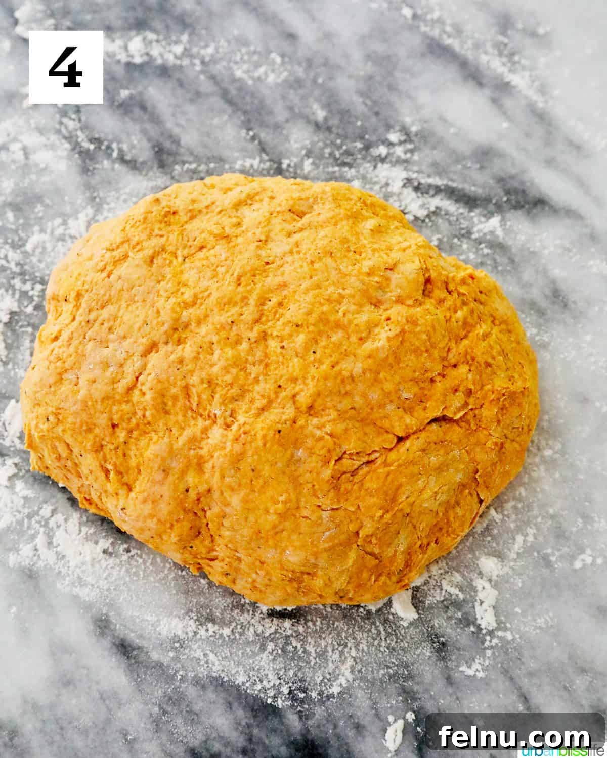A smooth, round ball of pumpkin gnocchi dough rests on a lightly floured marble countertop, indicating it's ready for the next stage of preparation after kneading.