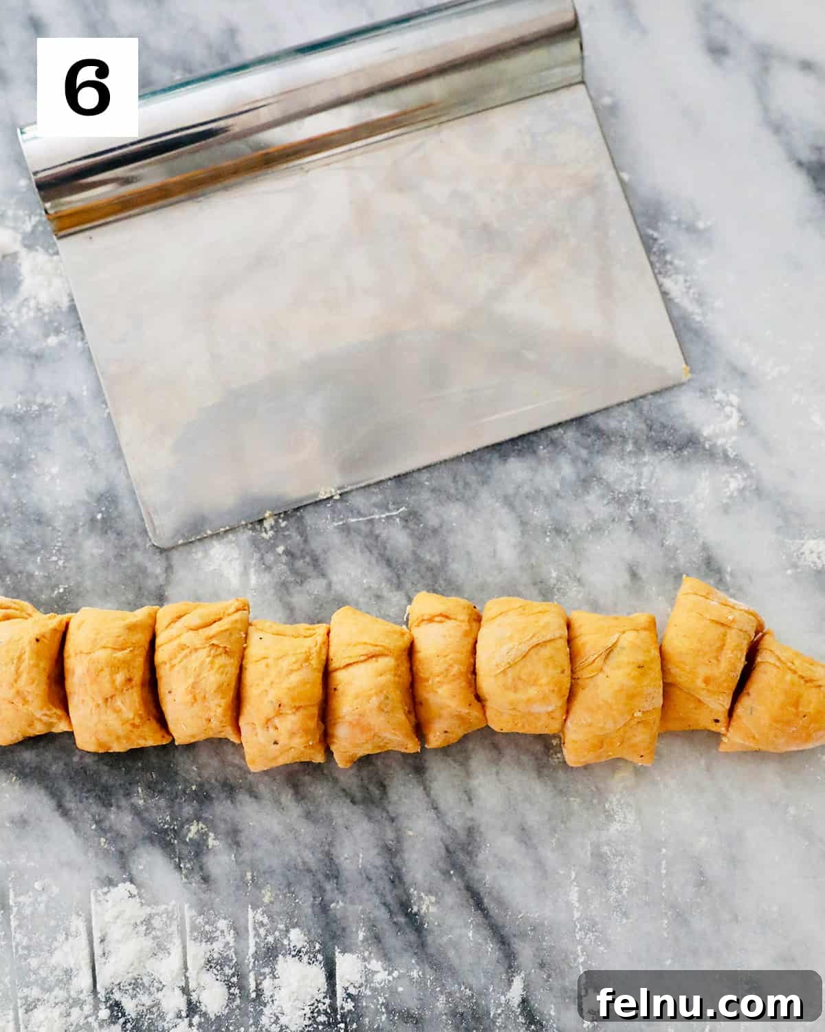 A pastry cutter precisely slices a rope of pumpkin gnocchi dough into small, uniform pieces on a marble countertop, demonstrating the portioning process.