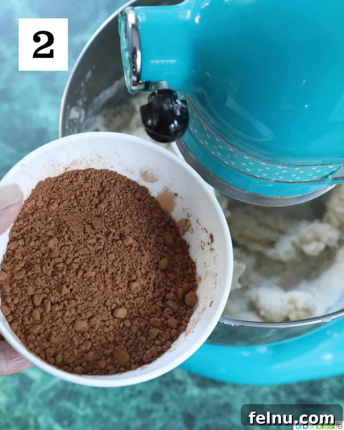 Effortless Decadent Chocolate Buttercream 6 Cocoa powder being carefully added to a blue stand mixer bowl containing creamed butter, the second step in making chocolate buttercream frosting.