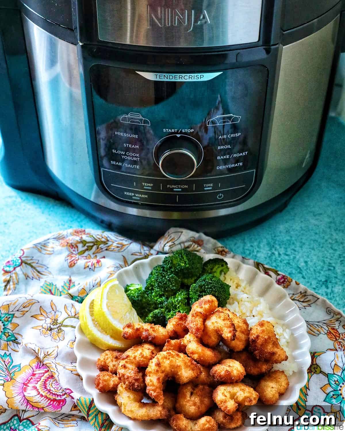 Crispy Air Fryer Popcorn Shrimp 10 A high-angle shot of a Ninja Foodi multicooker behind a plate of golden popcorn shrimp, steamed broccoli, and lemon wedges.