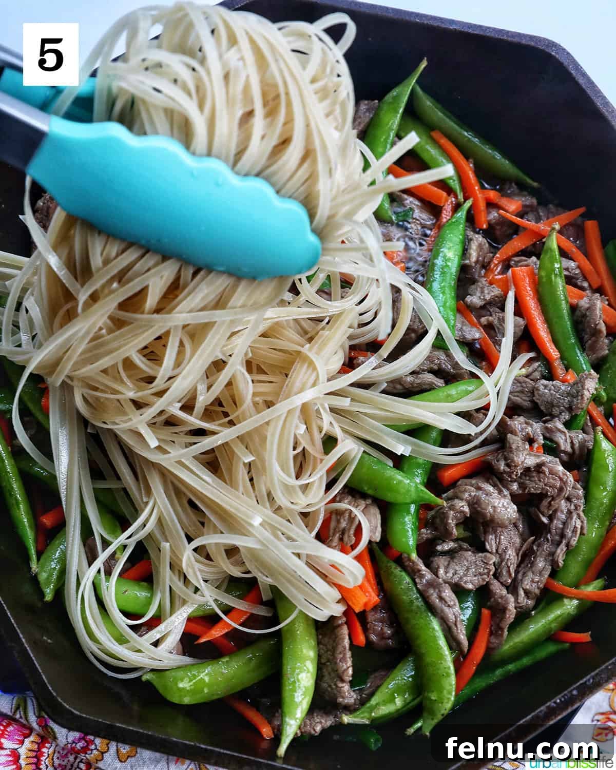 Beef Pad Thai 10 Blue tongs holding up beef pad thai noodles with vegetables in a cast iron skillet.