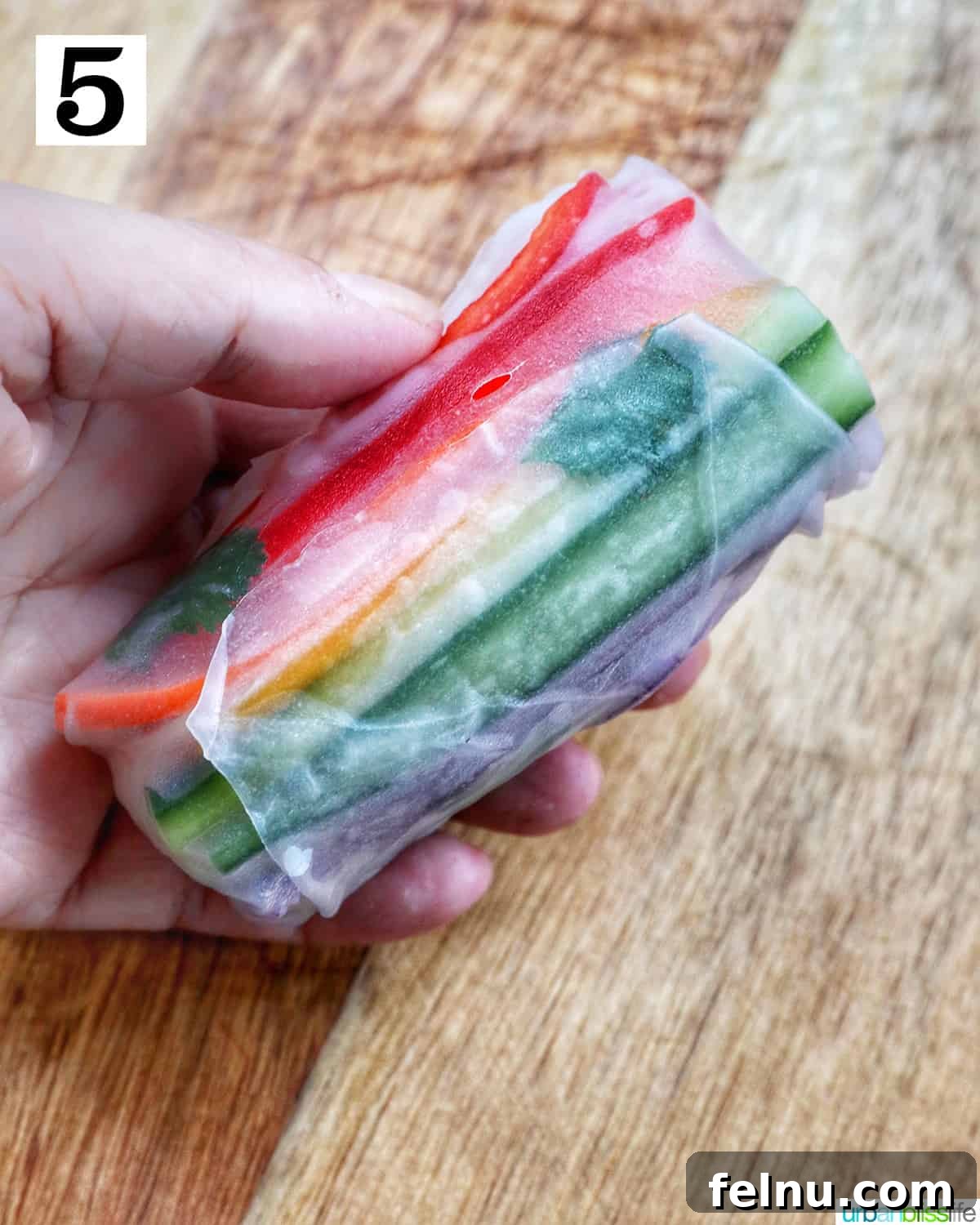 Vegan Delight: Fresh Summer Rolls 9 A hand holding a perfectly rolled vegan summer roll, filled with a colorful array of sliced vegetables, above a wooden cutting board.