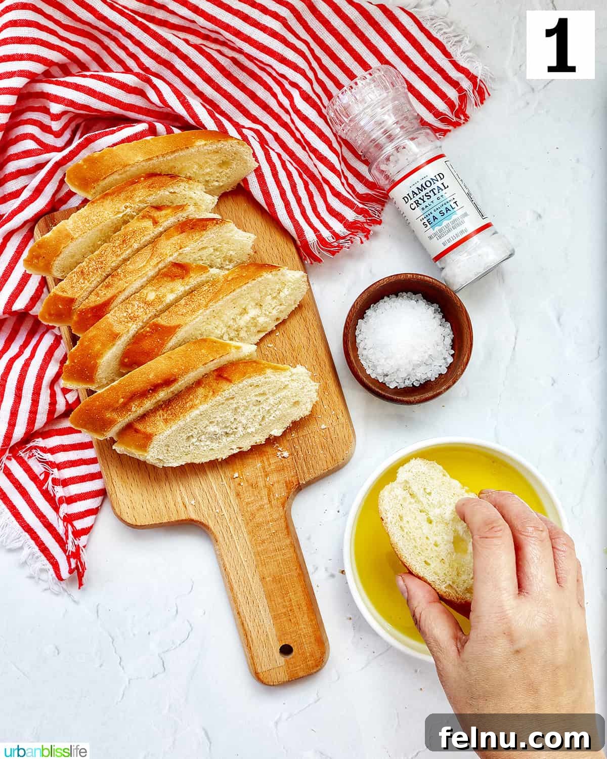 A hand dipping a baguette slice into a shallow bowl of olive oil on a wooden cutting board, with other baguette slices waiting.