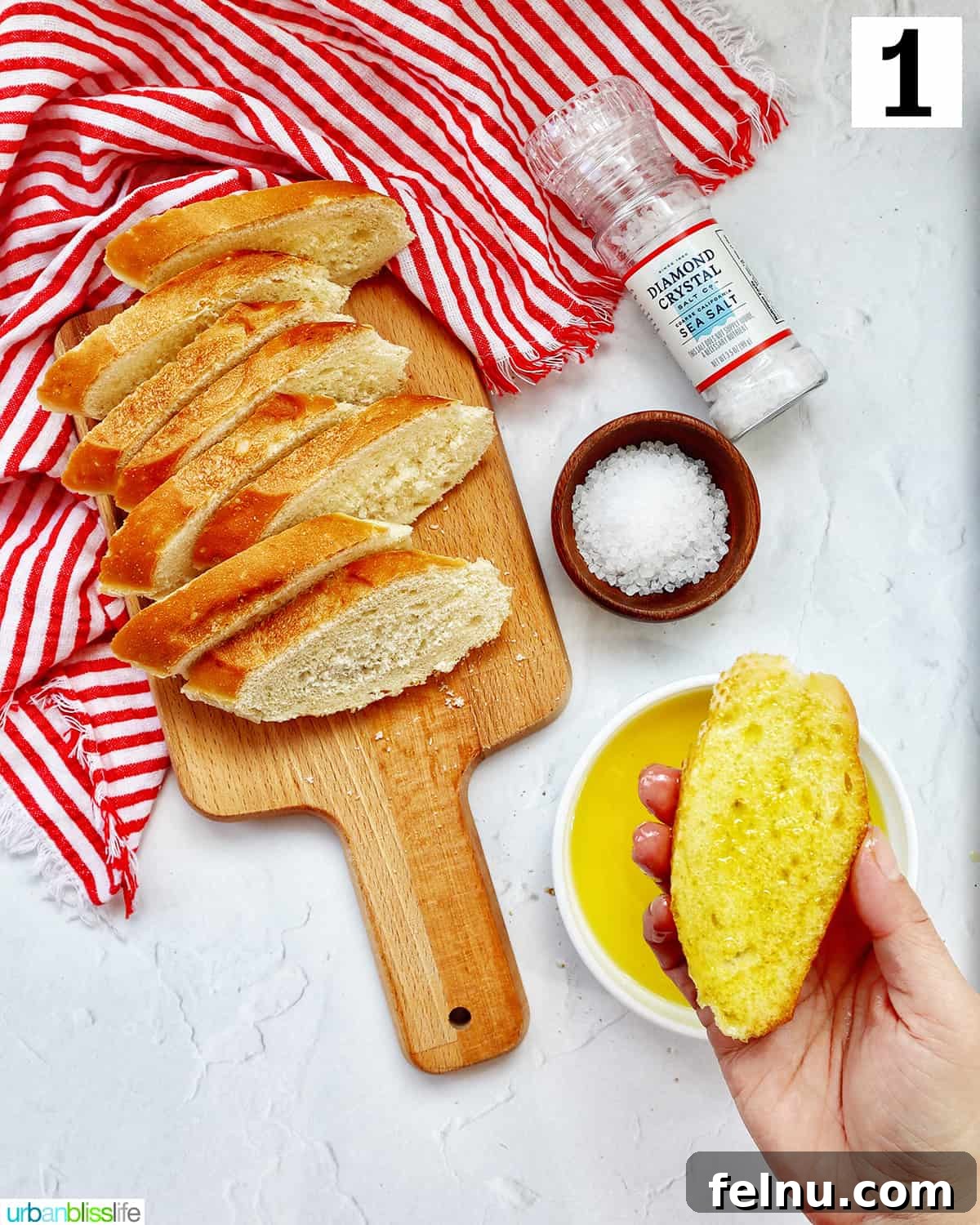 A baguette slice being coated with olive oil on one side, ready for baking, next to a bowl of olive oil.