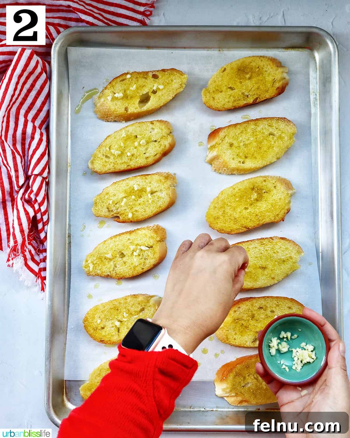 A hand sprinkling minced garlic evenly over olive oil-coated baguette slices arranged on a baking sheet lined with parchment paper.