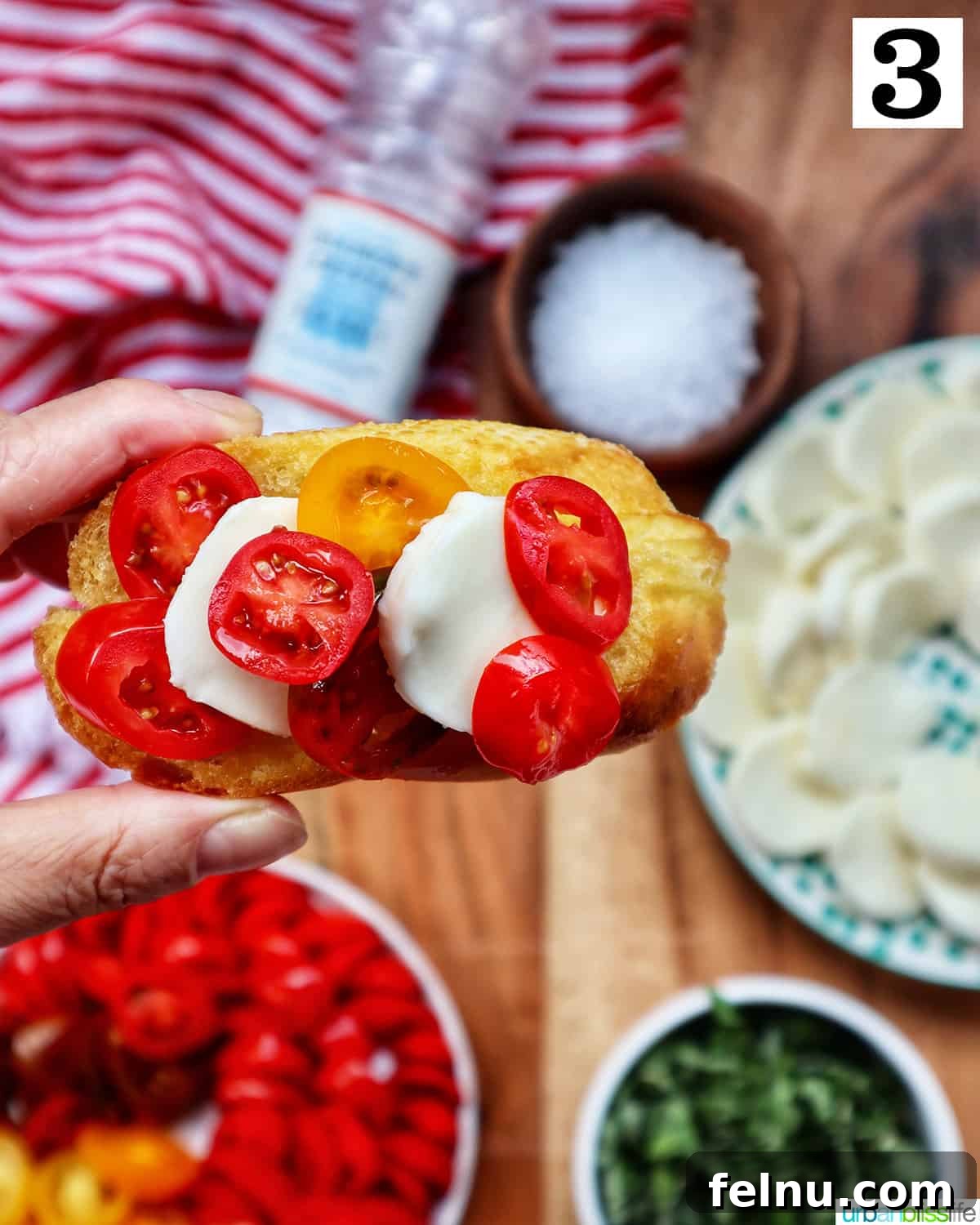 A hand holding a finished Caprese Crostini, showcasing the vibrant toppings of tomato, mozzarella, and basil. Background shows more prepared crostini.