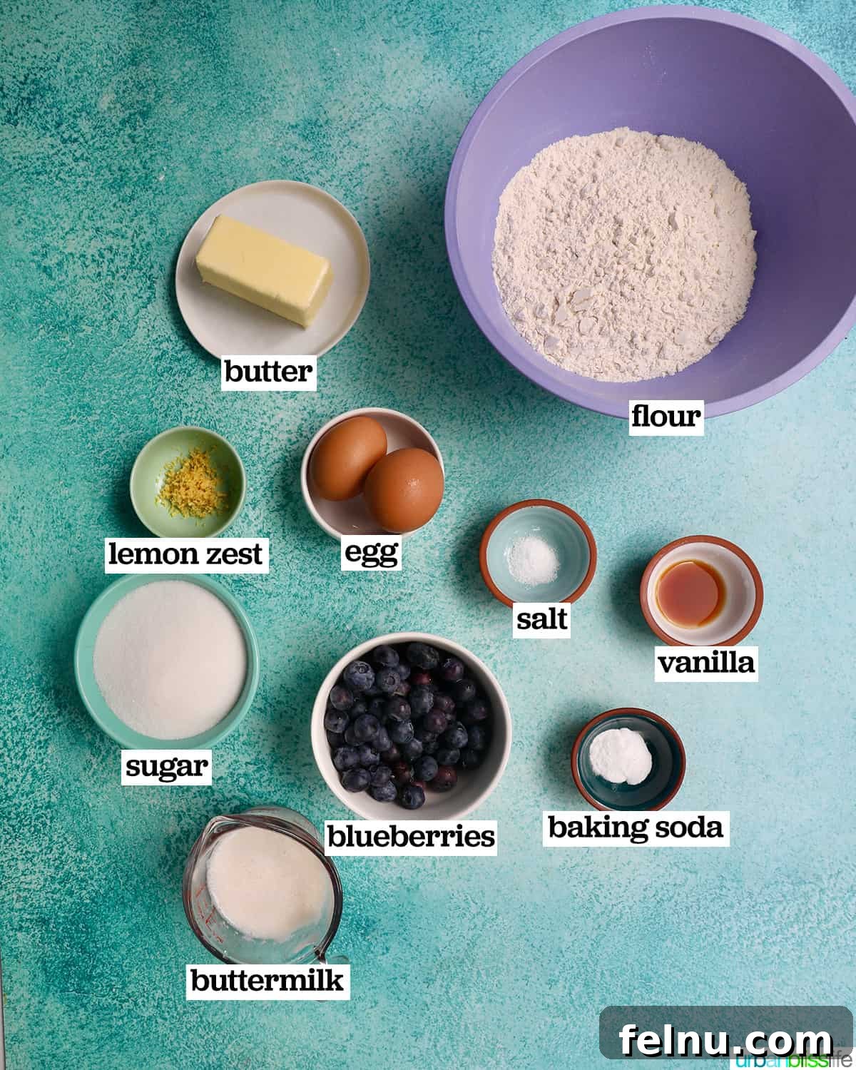Bowls of key ingredients for making lemon blueberry pound cake, including flour, sugar, lemon zest, and blueberries, set against a vibrant blue background.