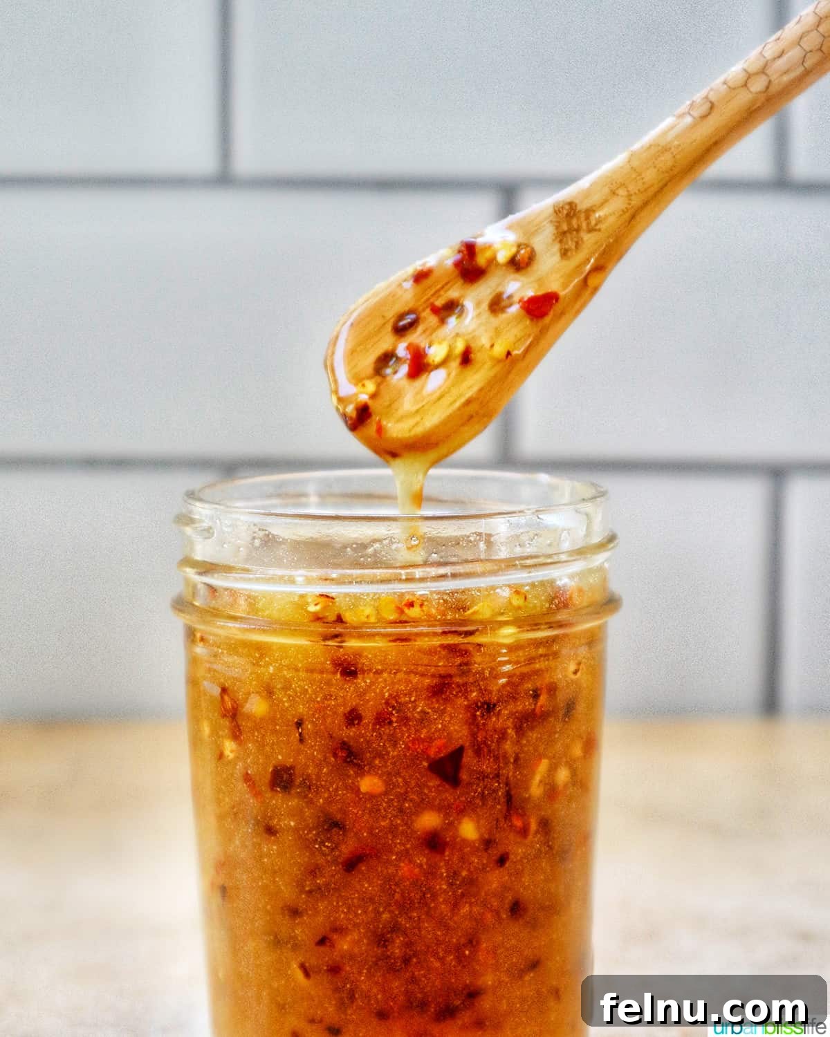 Hot Honey Sauce dripping from a wooden spoon into a mason jar, set against a subway tile kitchen backsplash.