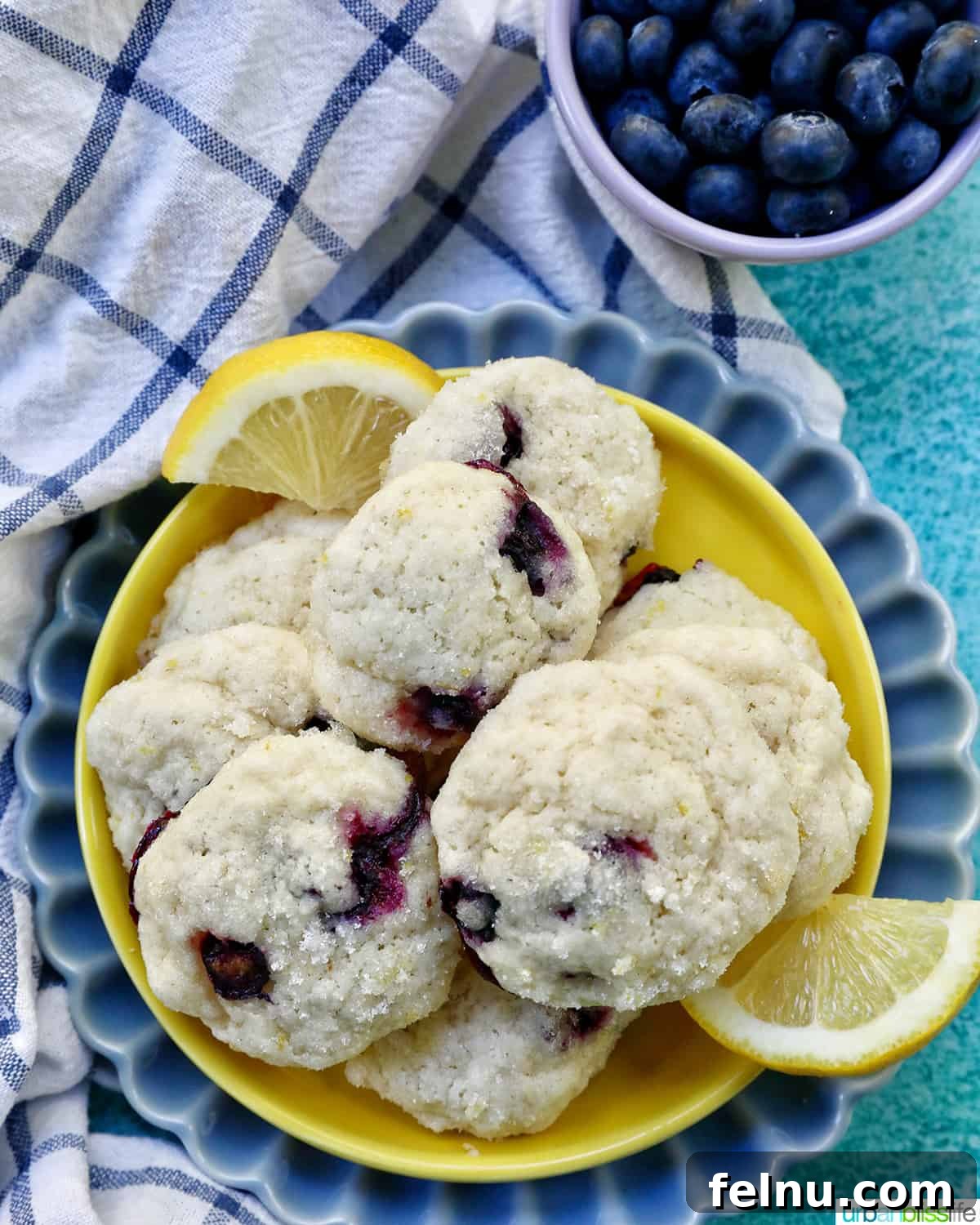 Lemon Blueberry Bliss Cookies 2 Lemon blueberry cookies on a yellow and blue plate with lemon slices, bowl of blueberries, and white and blue napkin.