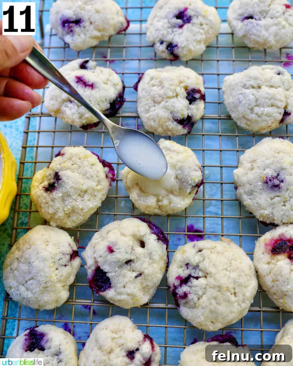 Lemon Blueberry Bliss Cookies 20 Spoon of lemon glaze over lemon blueberry cookies on a wire rack with white and blue napkin.