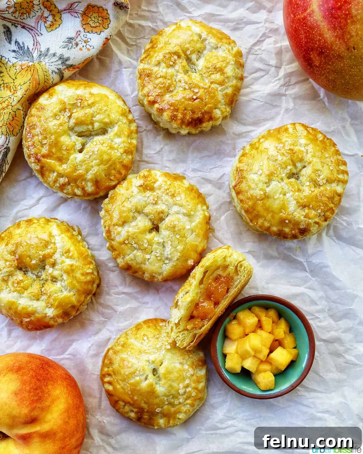 Sun-Kissed Peach Mango Pie 2 Several mini peach mango hand pies on crinkled parchment paper, with a bowl of diced peaches and mangoes in the background.