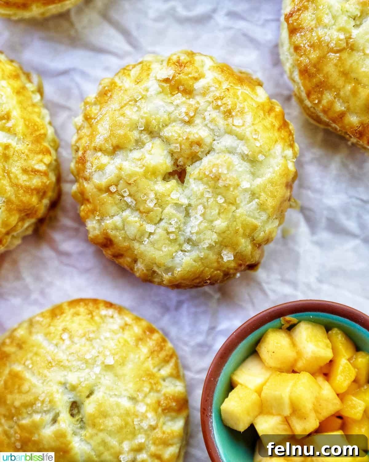 Sun-Kissed Peach Mango Pie 16 Several mini peach mango hand pies on crinkled parchment paper, with a bowl of diced peaches and mangoes in the background, showing a close-up of the golden crust.