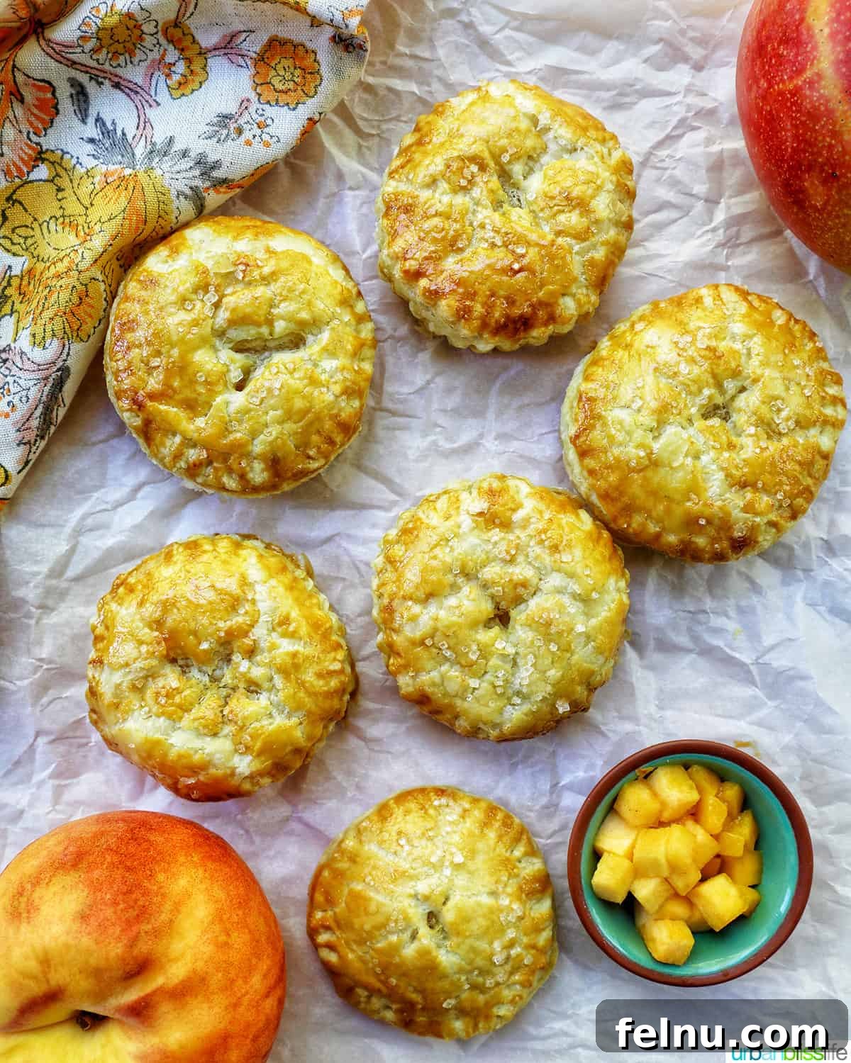 Sun-Kissed Peach Mango Pie 17 Several mini peach mango hand pies on crinkled parchment paper, with a bowl of diced peaches and mangoes from an overhead view.