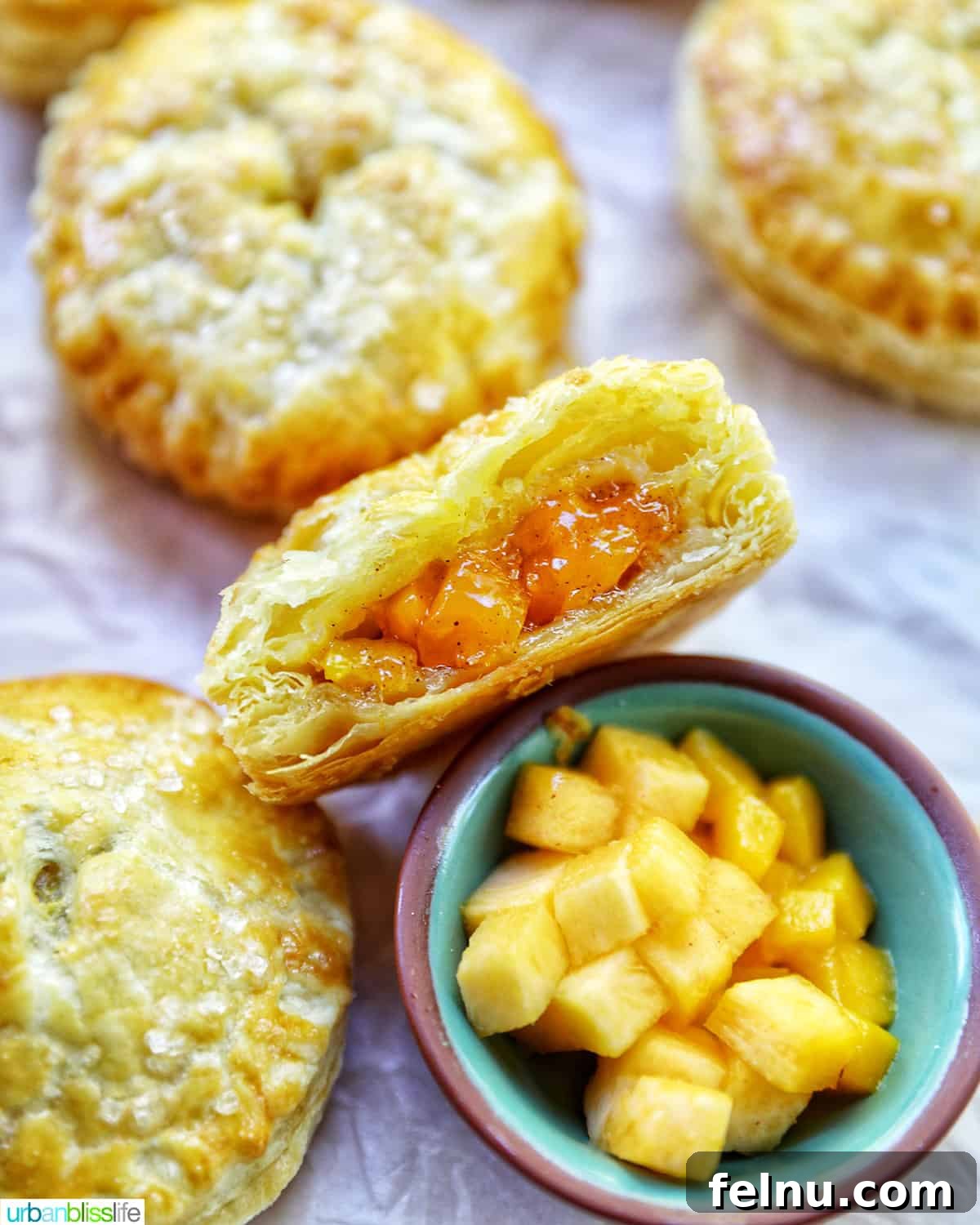 Sun-Kissed Peach Mango Pie 3 Several mini peach mango hand pies on crinkled parchment paper with a bowl of diced peaches and mangoes.