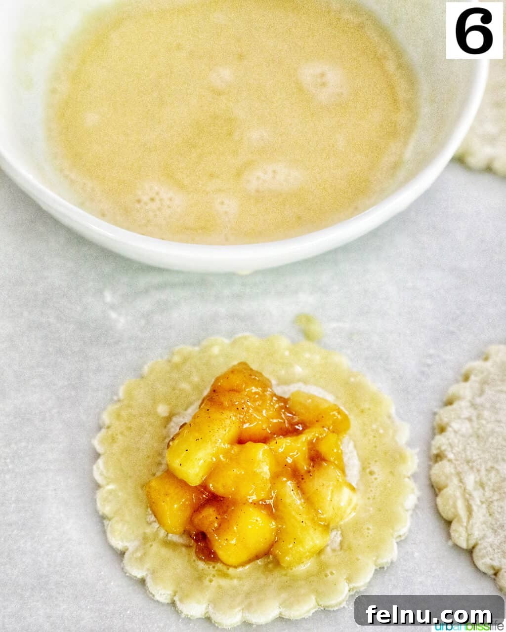 Sun-Kissed Peach Mango Pie 10 Egg wash in a bowl, positioned above a puff pastry round with diced peaches and mangoes on top, ready for brushing.