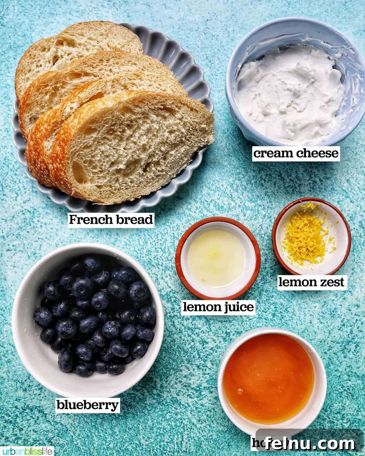 Blueberry Bliss Toast 4 Separate bowls of ingredients for blueberry toast: fresh blueberries, honey, lemon, cream cheese, and slices of bread on a blue table.