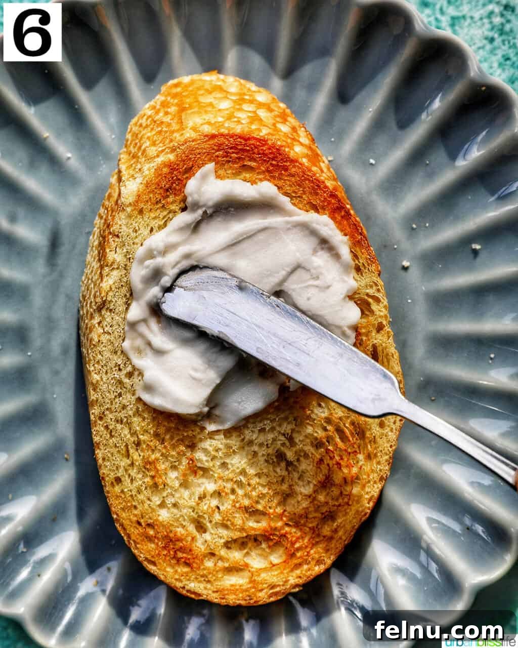 Blueberry Bliss Toast 10 Spreading smooth cream cheese evenly onto a slice of toasted bread on a blue plate.